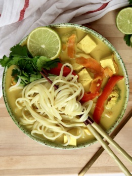 Large bowl with yellow curry broth inside with tofu, noodles, red bell pepper and lime to the side, with chopsticks through the noodles.