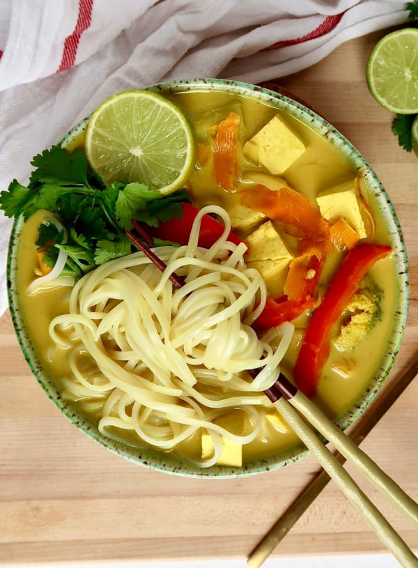 Large bowl with yellow curry broth inside with tofu, noodles, red bell pepper and lime to the side, with chopsticks through the noodles.