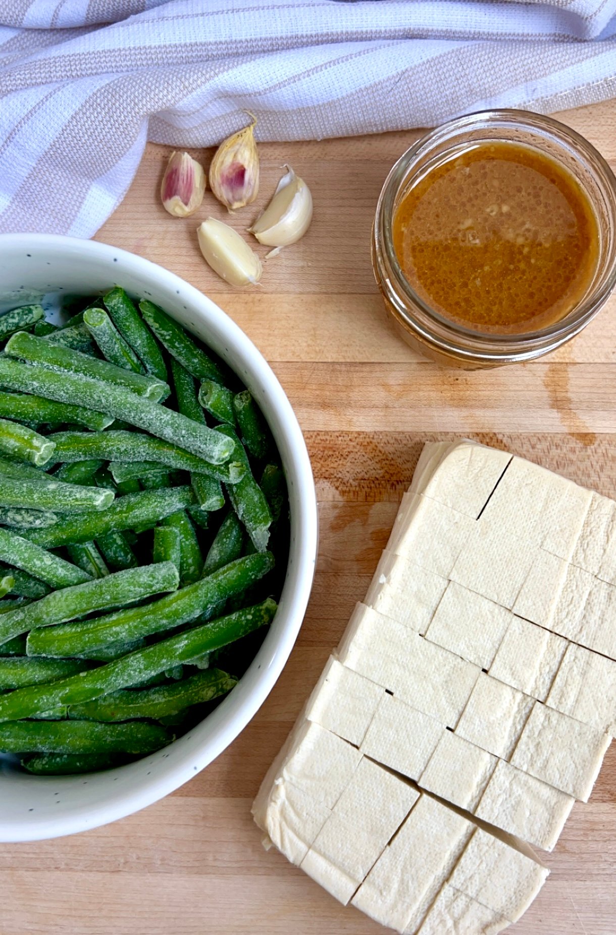 Cutting board with cubed tofu, frozen green beans, sauce in a bowl and garlic cloves.