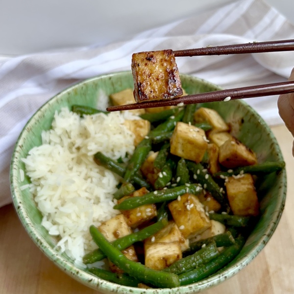 Bowl of tofu with green beans and rice on the side and a chopstick holding one piece of tofu