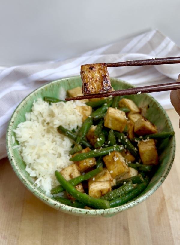 Bowl of tofu with green beans and rice on the side and a chopstick holding one piece of tofu