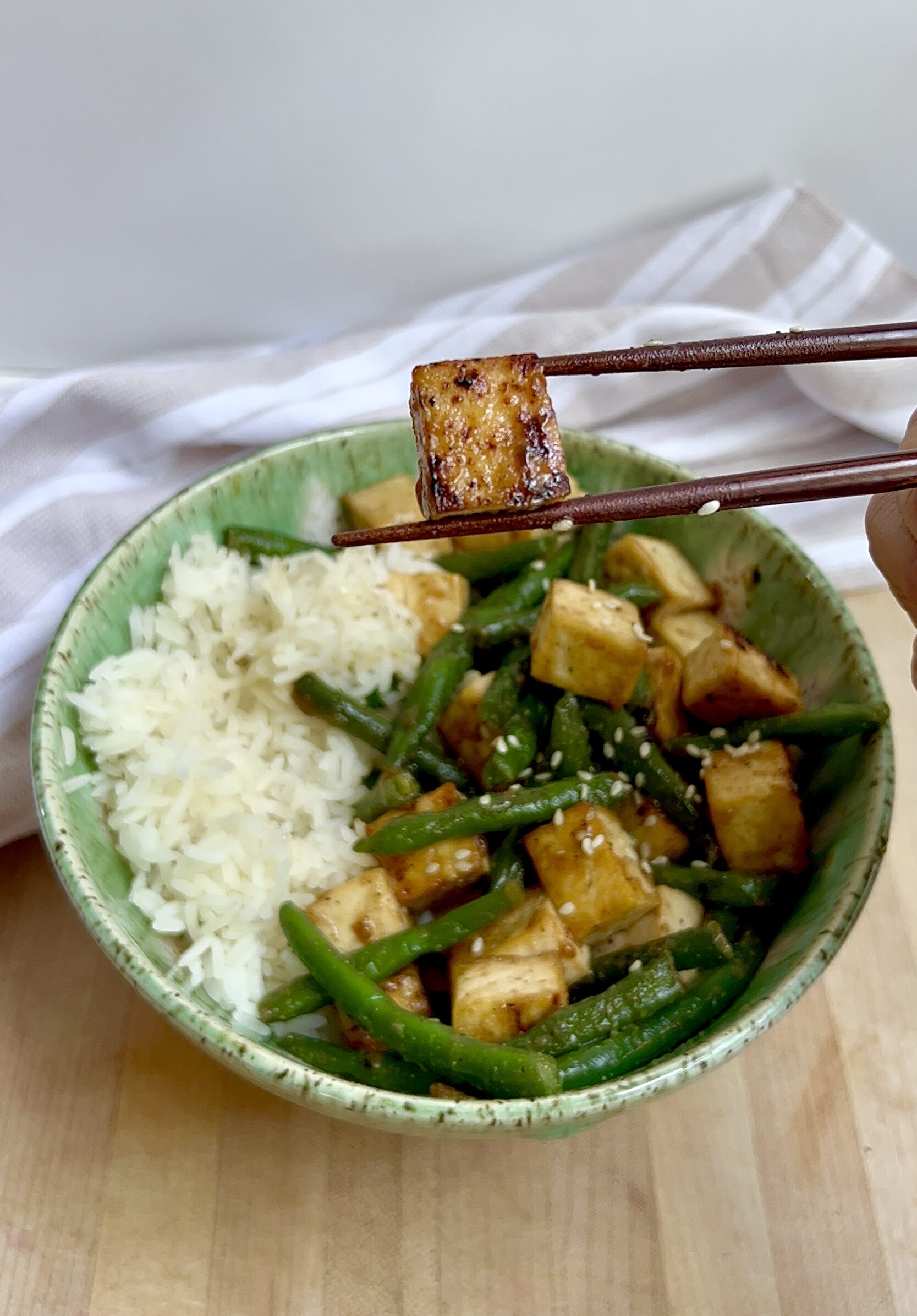Bowl of tofu with green beans and rice on the side and a chopstick holding one piece of tofu