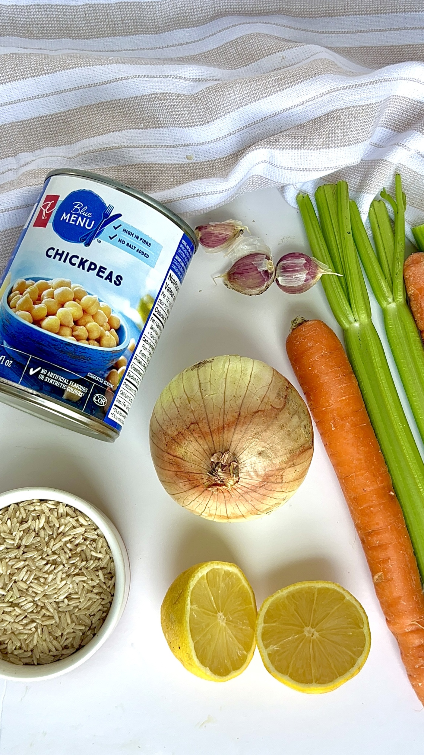 White background with canned chickpeas, carrots, onion, garlic, uncooked brown rice and celery on it.