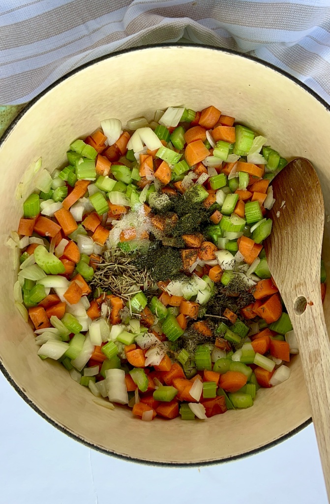 Large pot with celery, onion and carrots with spices and a wooden spoon stirring.