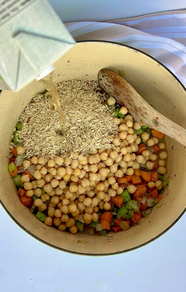 Large pot with chickpeas, uncooked rice and diced veggies inside and veggie stock being poured in.