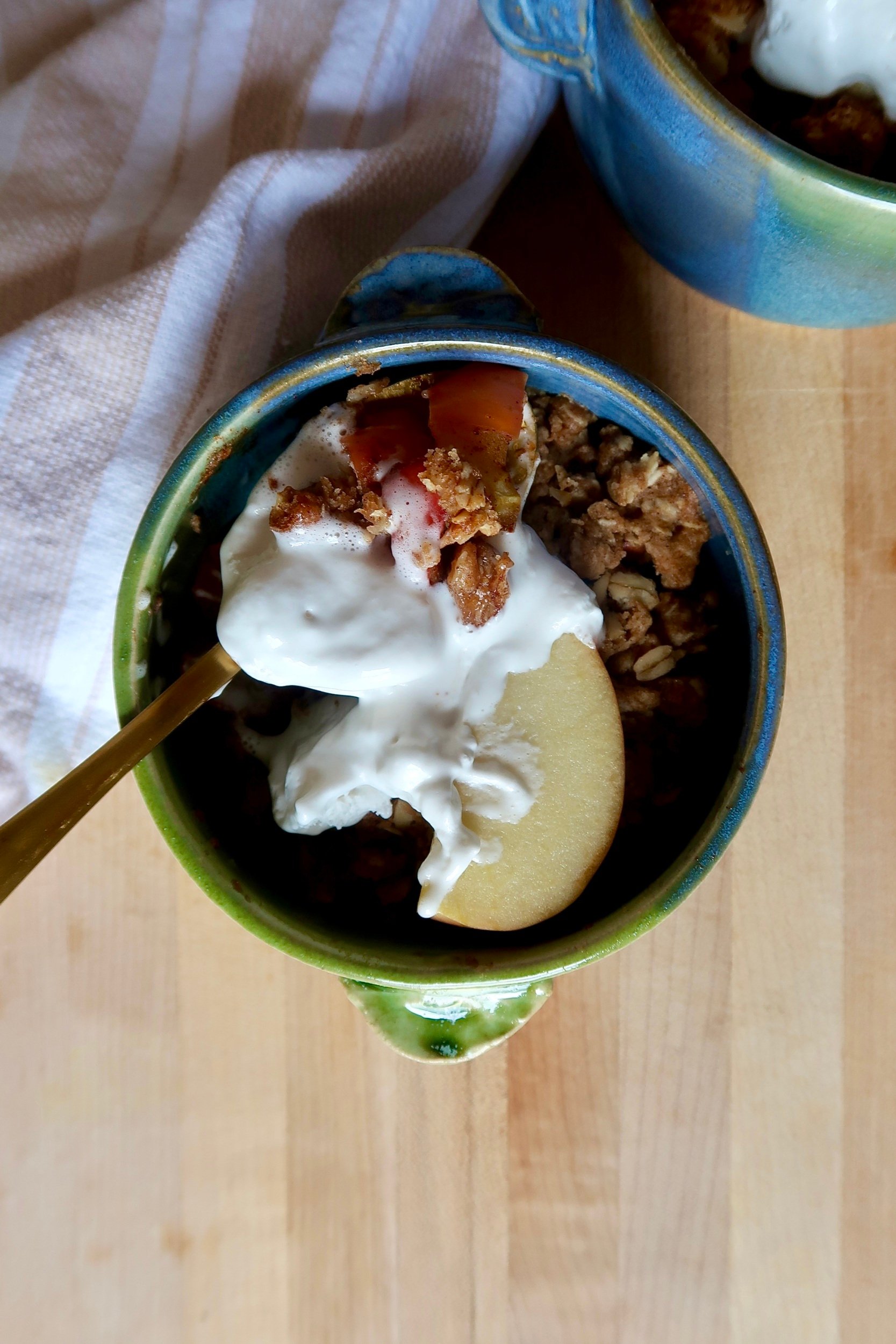 Blue and green ramekin with apple crisp inside and whipped cream with a spoon holding some.