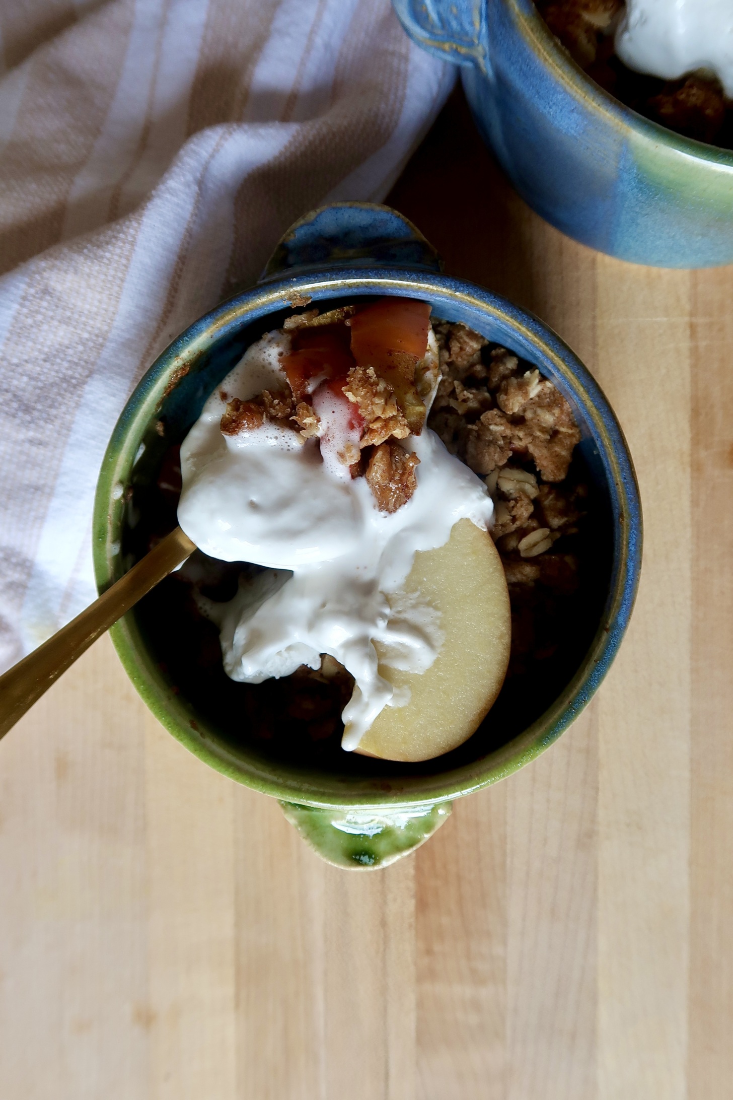 Blue and green ramekin with apple crisp inside and whipped cream with a spoon holding some.