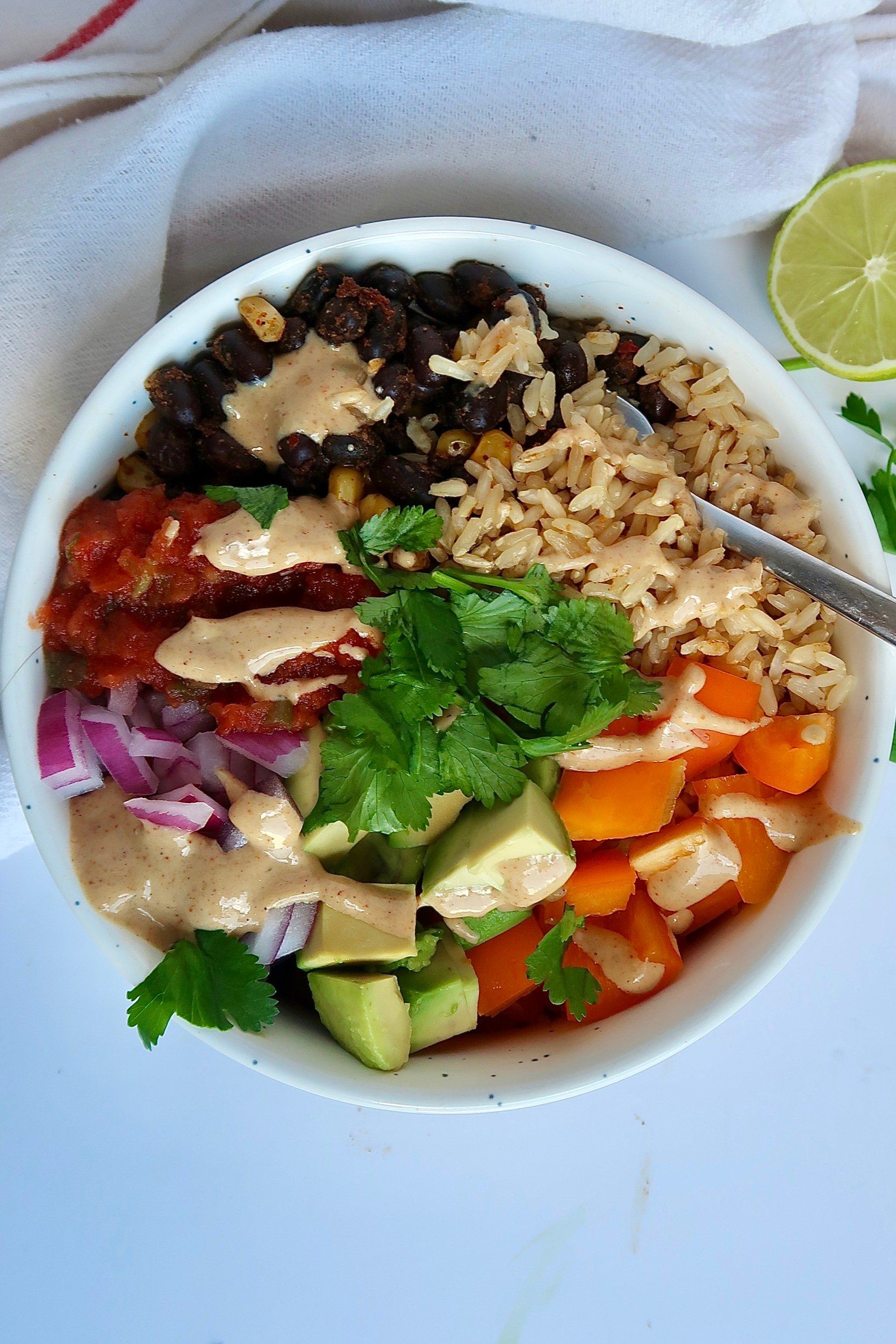 White bowl with spiced black beans and veggies inside as well as brown rice, salsa, and a creamy sauce on top