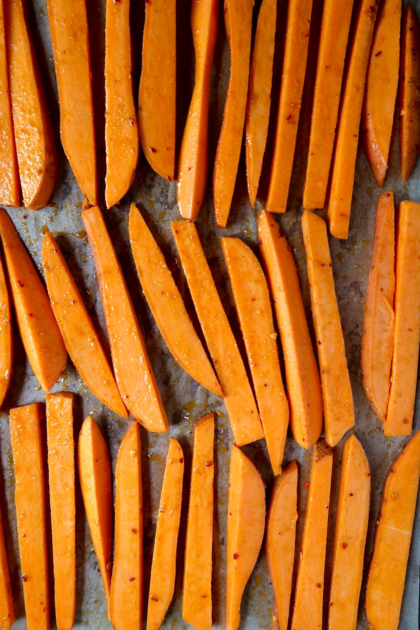 Sliced sweet potato fries covered in olive oil and spices on a sheet pan.