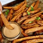 Cooked sweet potato fries on a sheet pan and a creamy sauce in a small bowl on the side, with a fry in the dipping sauce.