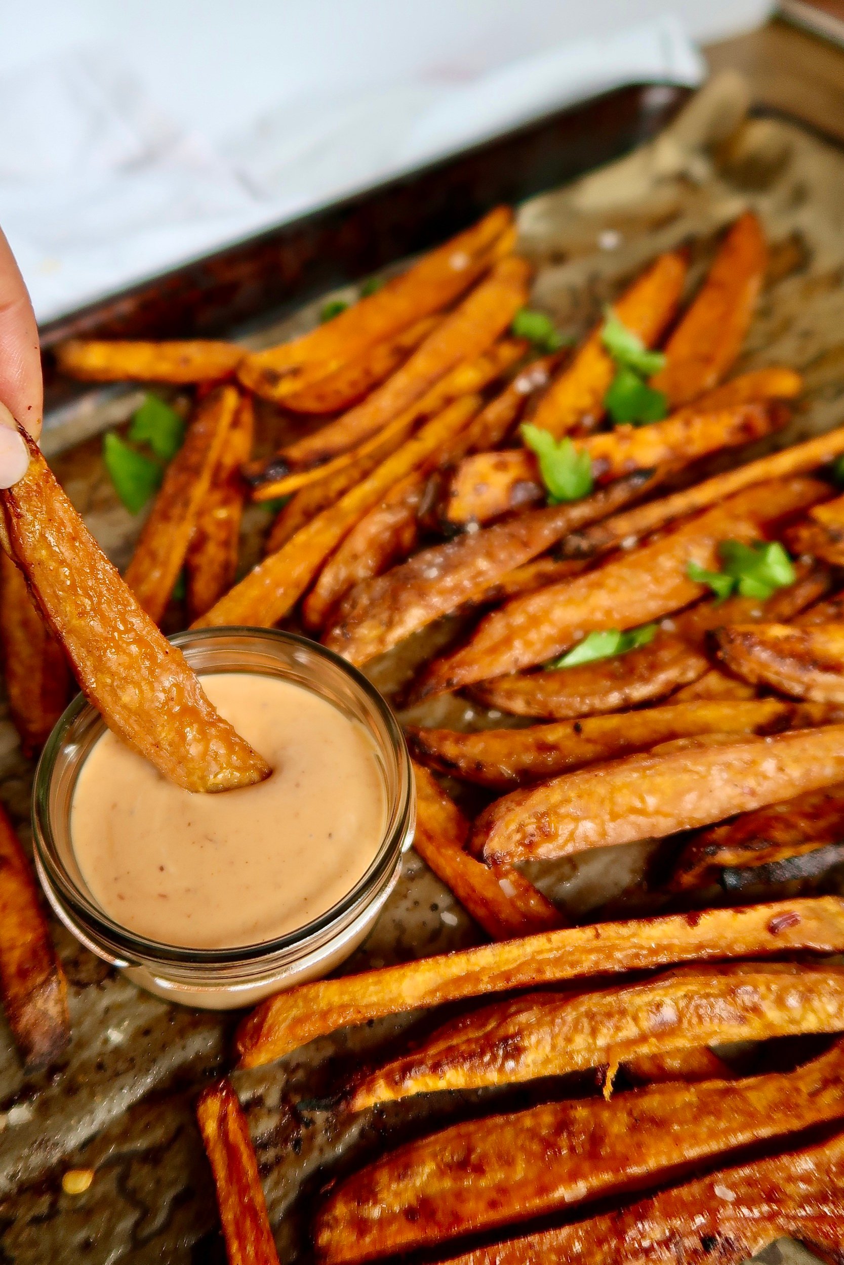 Cooked sweet potato fries on a sheet pan and a creamy sauce in a small bowl on the side, with a fry in the dipping sauce.