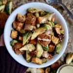 Hand holding a white bowl with diced sweet potato, brussel sprouts and vegan sausage inside with a sauce drizzled on top.