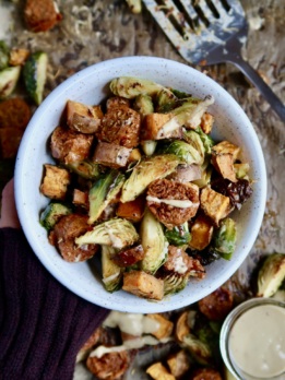 Hand holding a white bowl with diced sweet potato, brussel sprouts and vegan sausage inside with a sauce drizzled on top.