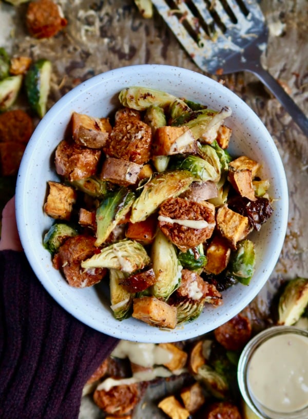 Hand holding a white bowl with diced sweet potato, brussel sprouts and vegan sausage inside with a sauce drizzled on top.