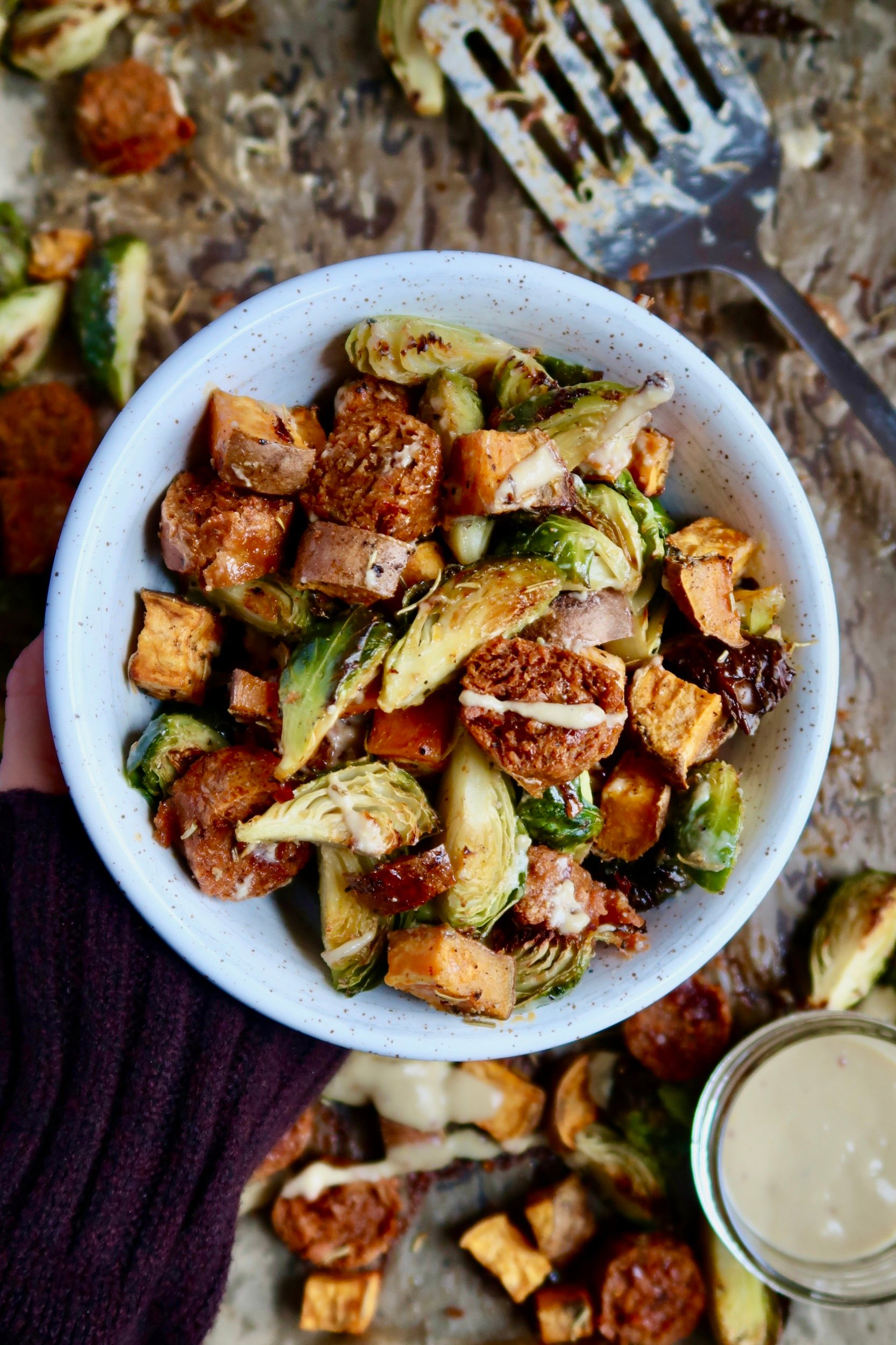 Hand holding a white bowl with diced sweet potato, brussel sprouts and vegan sausage inside with a sauce drizzled on top.