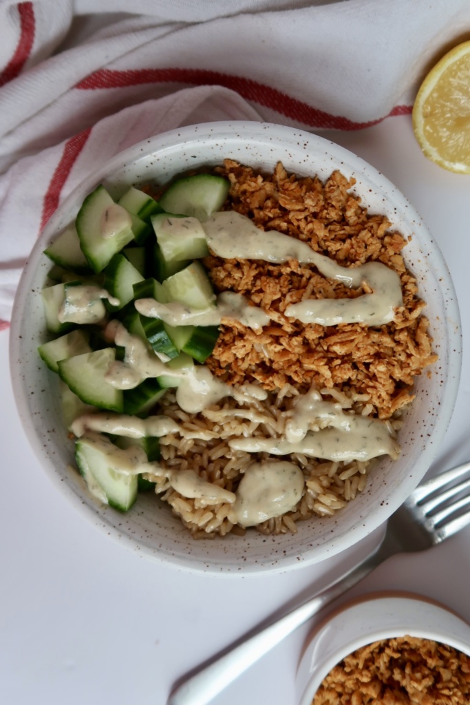 Shredded tofu in a bowl with cucumber and rice with tahini sauce drizzled on top.