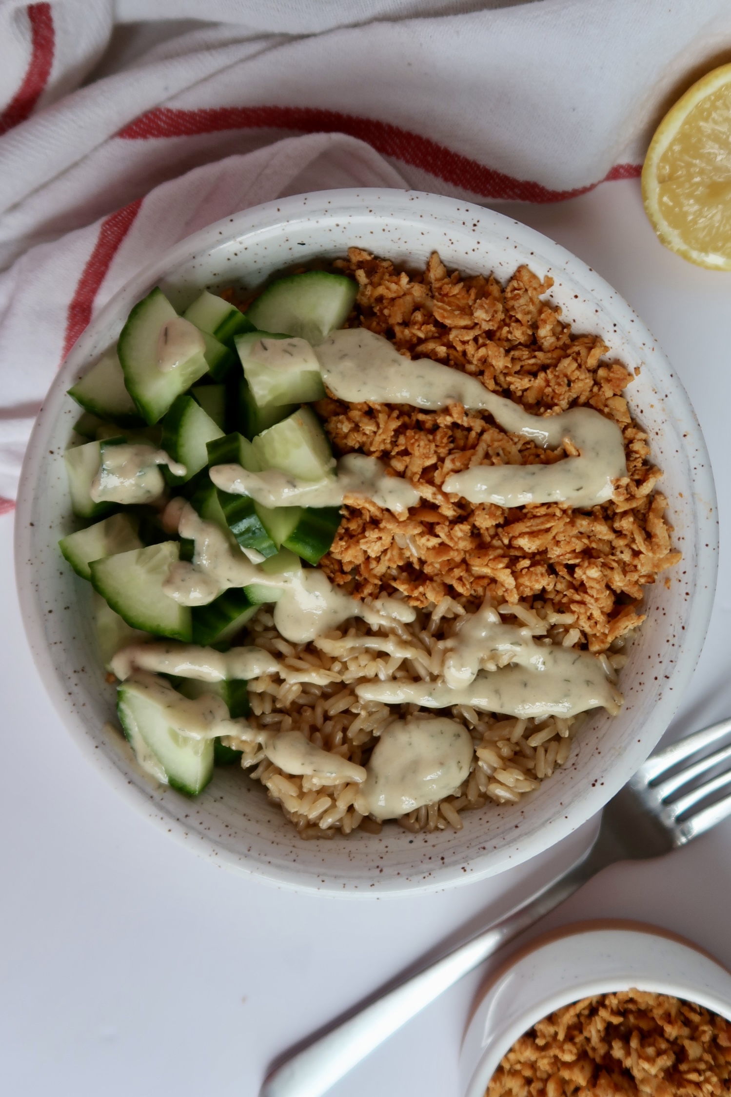 Shredded tofu in a bowl with cucumber and rice with tahini sauce drizzled on top.