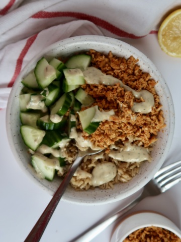Shredded tofu in a bowl with cucumber and rice with tahini sauce drizzled on top and a spoon scooping some out.