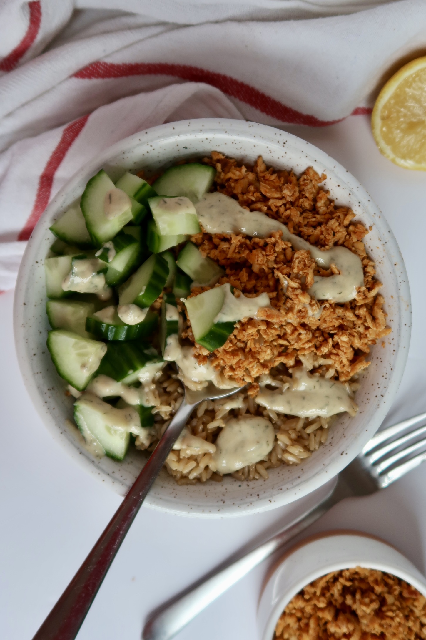 Shredded tofu in a bowl with cucumber and rice with tahini sauce drizzled on top and a spoon scooping some out.