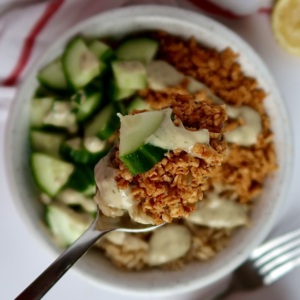Shredded tofu in a bowl with cucumber and rice with tahini sauce drizzled on top and a spoon scooping some out.