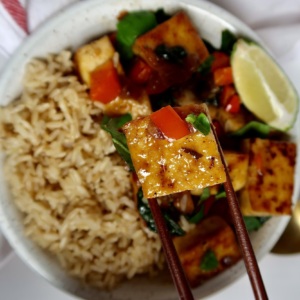 Cubed basil tofu in a bowl with brown rice on the side and a lime wedge with chopsticks holding up the tofu.