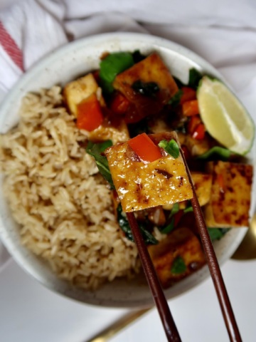 Cubed basil tofu in a bowl with brown rice on the side and a lime wedge with chopsticks holding up the tofu.