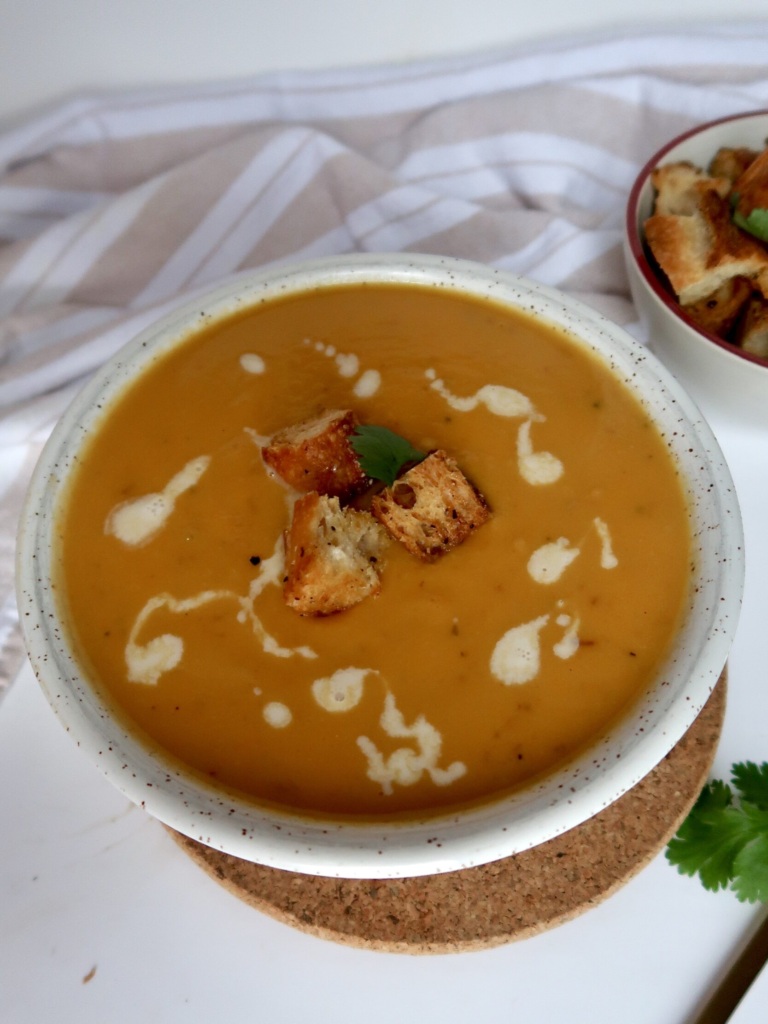 Side angle of Blended sweet potato soup in a white bowl with croutons on top and coconut milk drizzled inside.