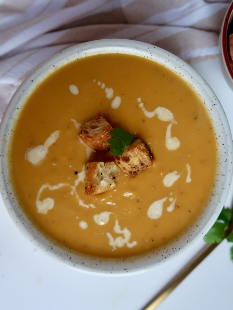 Blended sweet potato soup in a white bowl with croutons on top and coconut milk drizzled inside.