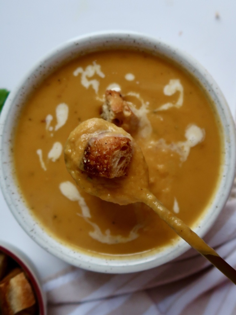 Blended sweet potato soup in a white bowl with croutons on top and coconut milk drizzled inside and a spoon with soup and a crouton.