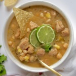 White bowl with creamy base and soy chunks with pieces of corn and beans with lime slices and cilantro on top and a spoon and corn chip in the bowl.