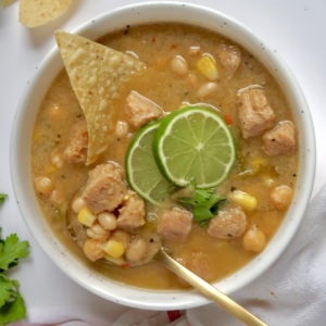 White bowl with creamy base and soy chunks with pieces of corn and beans with lime slices and cilantro on top and a spoon and corn chip in the bowl.