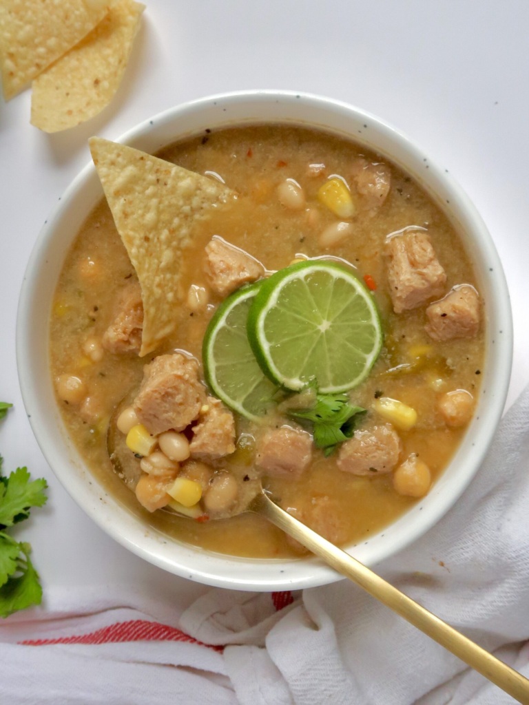 White bowl with creamy base and soy chunks with pieces of corn and beans with lime slices and cilantro on top and a spoon and corn chip in the bowl.