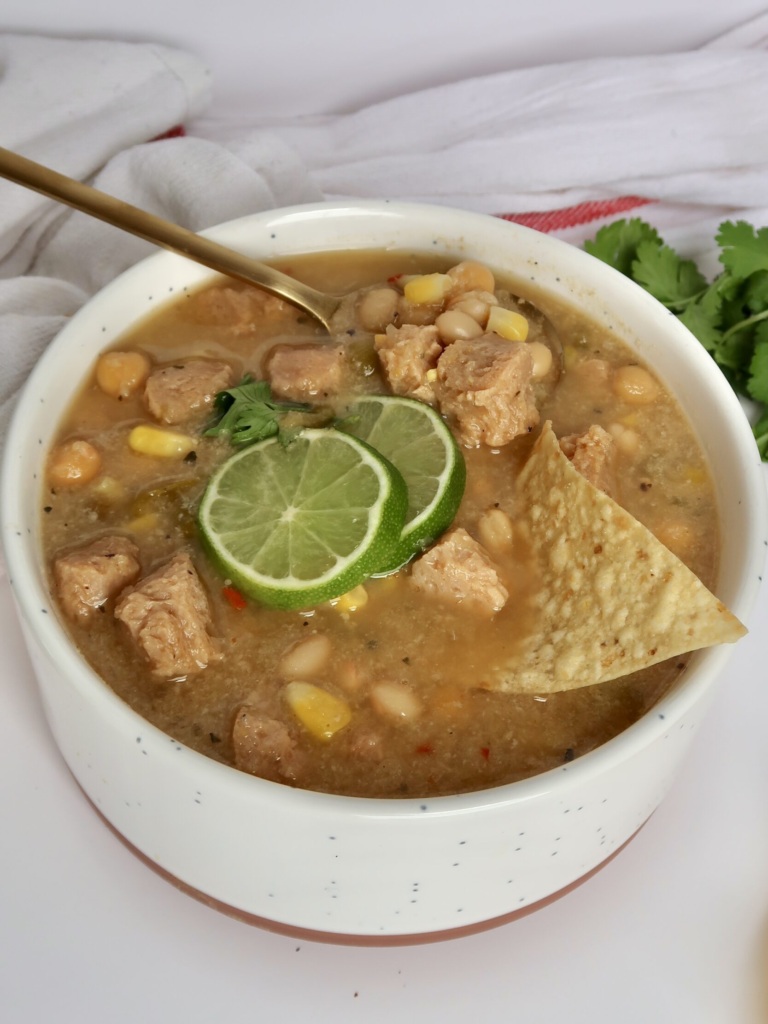 White bowl with creamy base and soy chunks with pieces of corn and beans with lime slices and cilantro on top and a spoon and corn chip in the bowl.