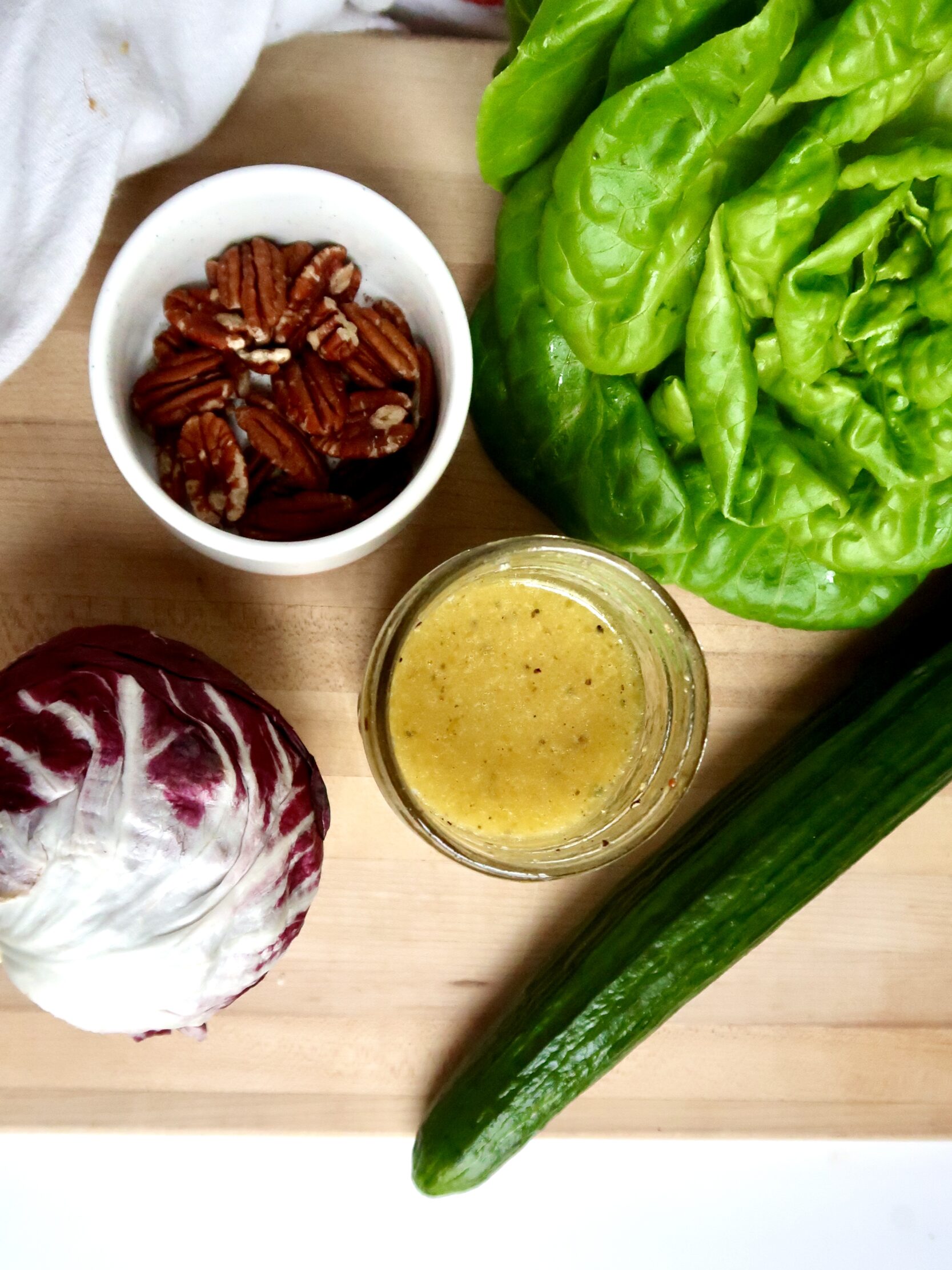 Cutting board with radicchio, butter lettuce, pecans, cucumbers and salad dressing.