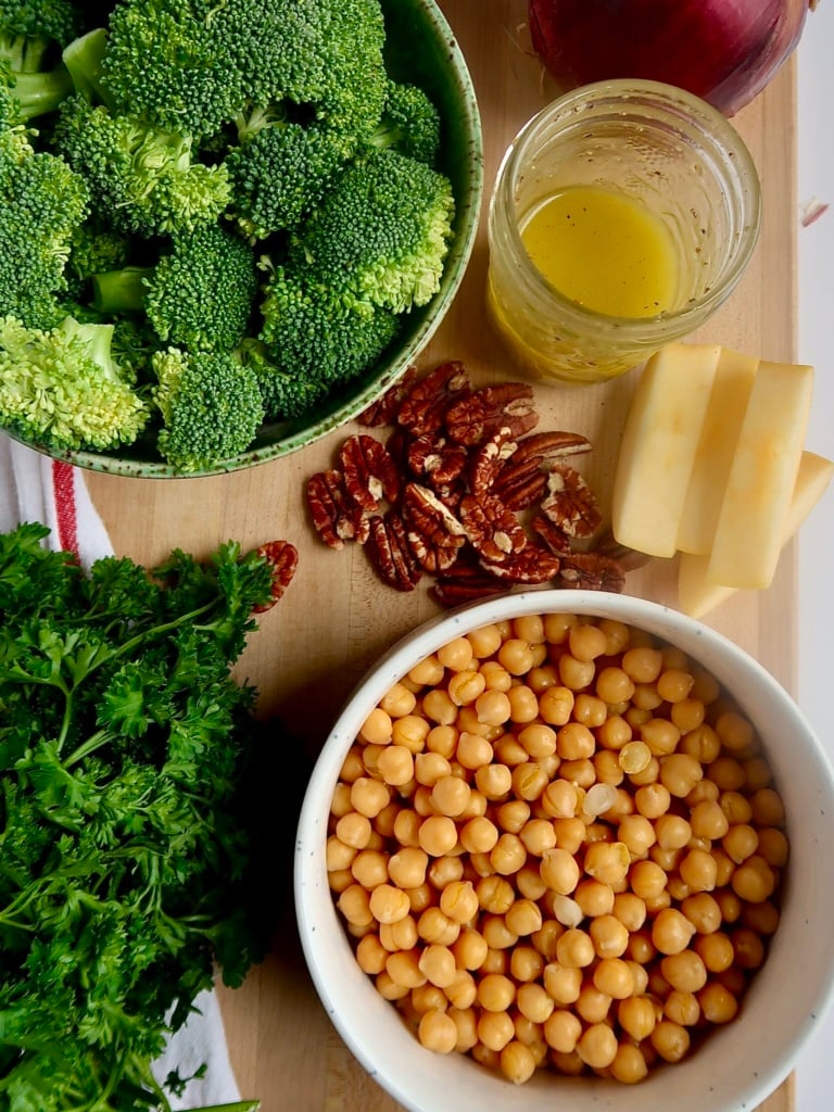 Chickpeas, broccoli, pecans, parsley, apple slices and vinaigrette in separate bowls on a cutting board.