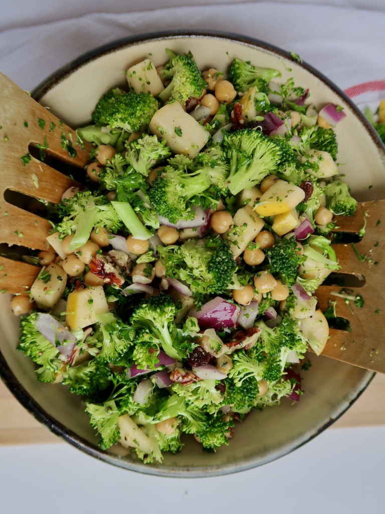 Bowl of broccoli, apple, red onion, chickpeas with salad tongs inside.