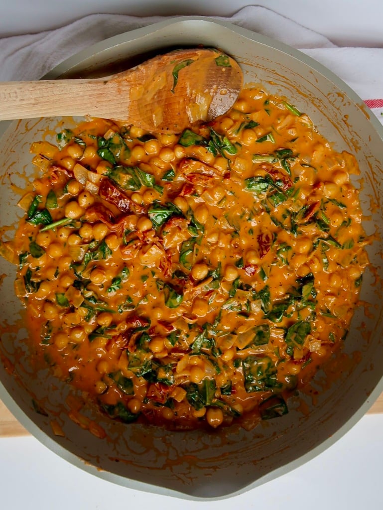 Pan with chickpeas mixed with tomato paste and coconut milk, sundried tomatoes and wilted spinach with a wooden spoon in the pan.