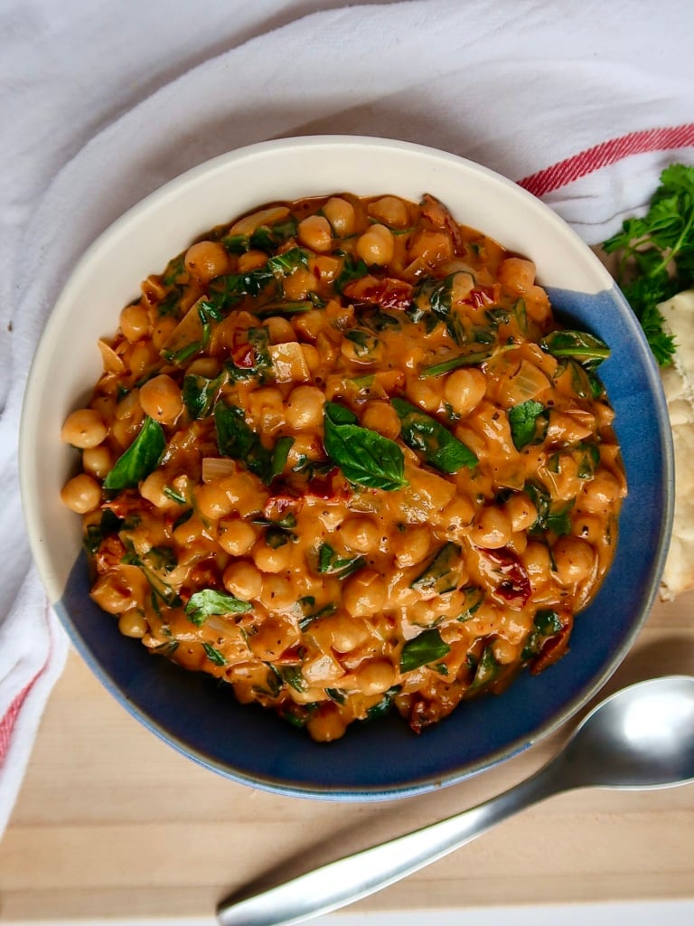 White and blue bowl with breasted chickpeas in a coconut and tomato paste mixture with spinach and basil in it as well.