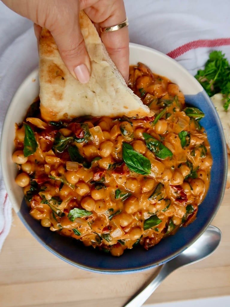 White and blue bowl with breasted chickpeas in a coconut and tomato paste mixture with spinach and basil, with a hang dipping a piece of naan.