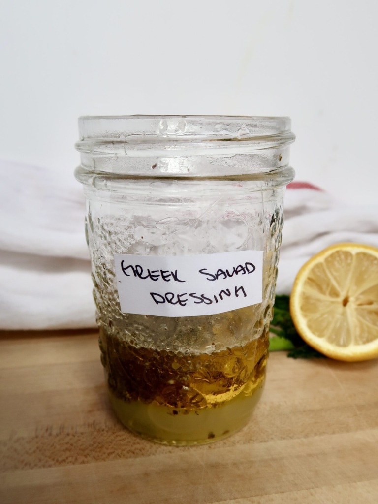 Jar with olive oil, lemon juice and herbs and a lemon in the background and labeled 'Greek Salad Dressing'