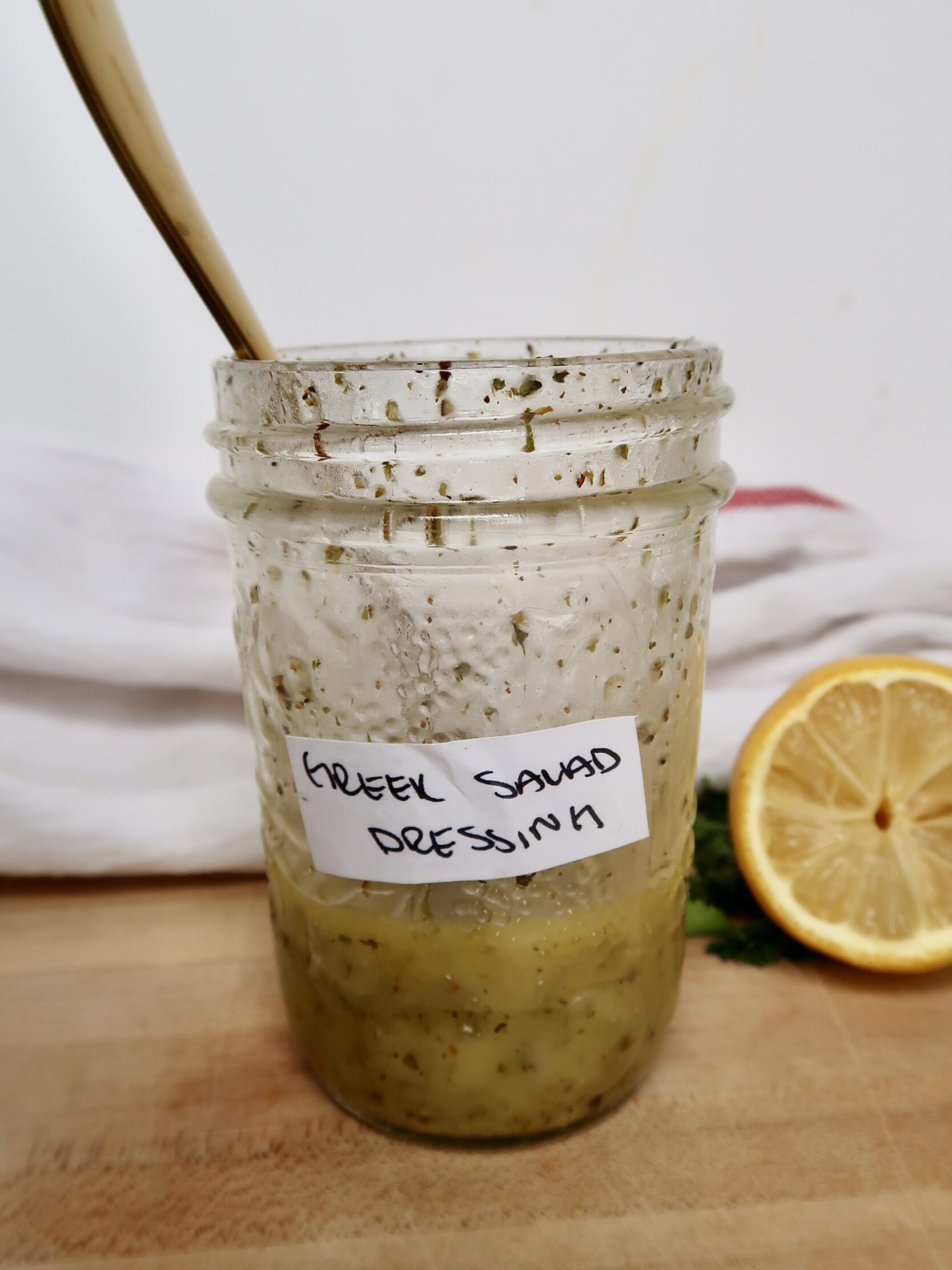 Jar labaled 'Greek Salad Dressing' with a spoon inside and a salad dressing with herbs.