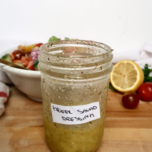 Jar with a salad dressing inside labeled 'Greek Salad Dressing', with a bowl of green salad and a lemon in the background.