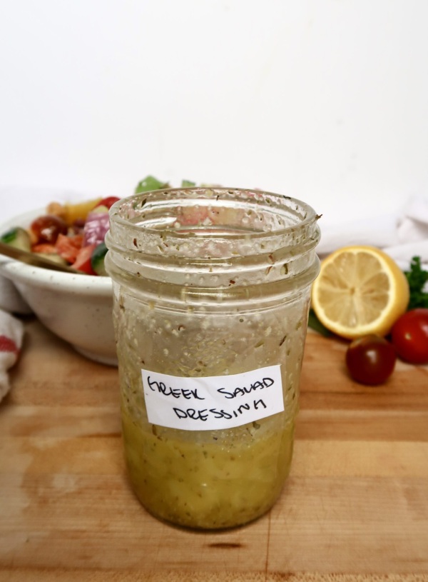 Jar with a salad dressing inside labeled 'Greek Salad Dressing', with a bowl of green salad and a lemon in the background.
