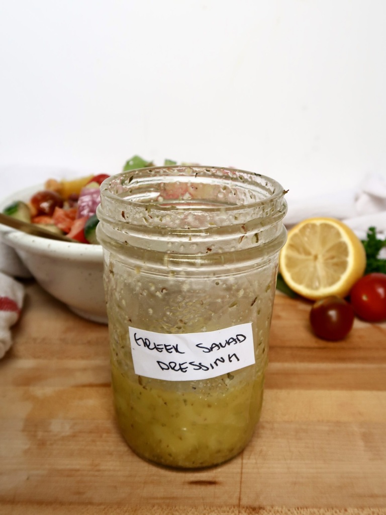 Jar with a salad dressing inside labeled 'Greek Salad Dressing', with a bowl of green salad and a lemon in the background.