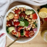 White bowl with chickpeas, tomatoes, cucumber, bell pepper inside with a jar labeled 'Greek salad dressing' and a lemon on the side.