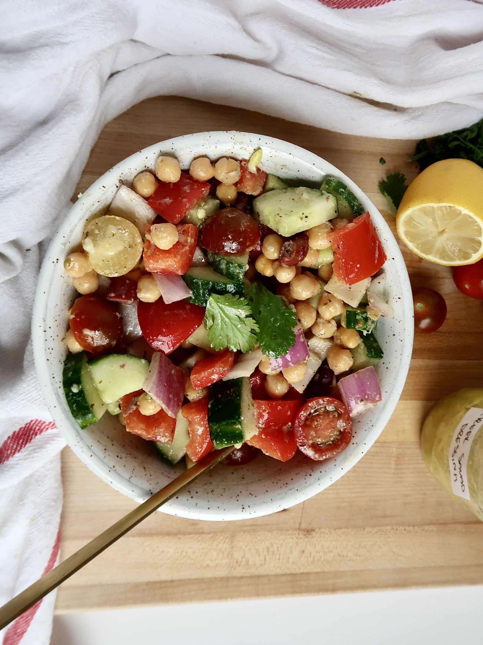 White bowl with chickpeas, tomatoes, cucumber, bell pepper inside with a jar labeled 'Greek salad dressing' and a lemon on the side.