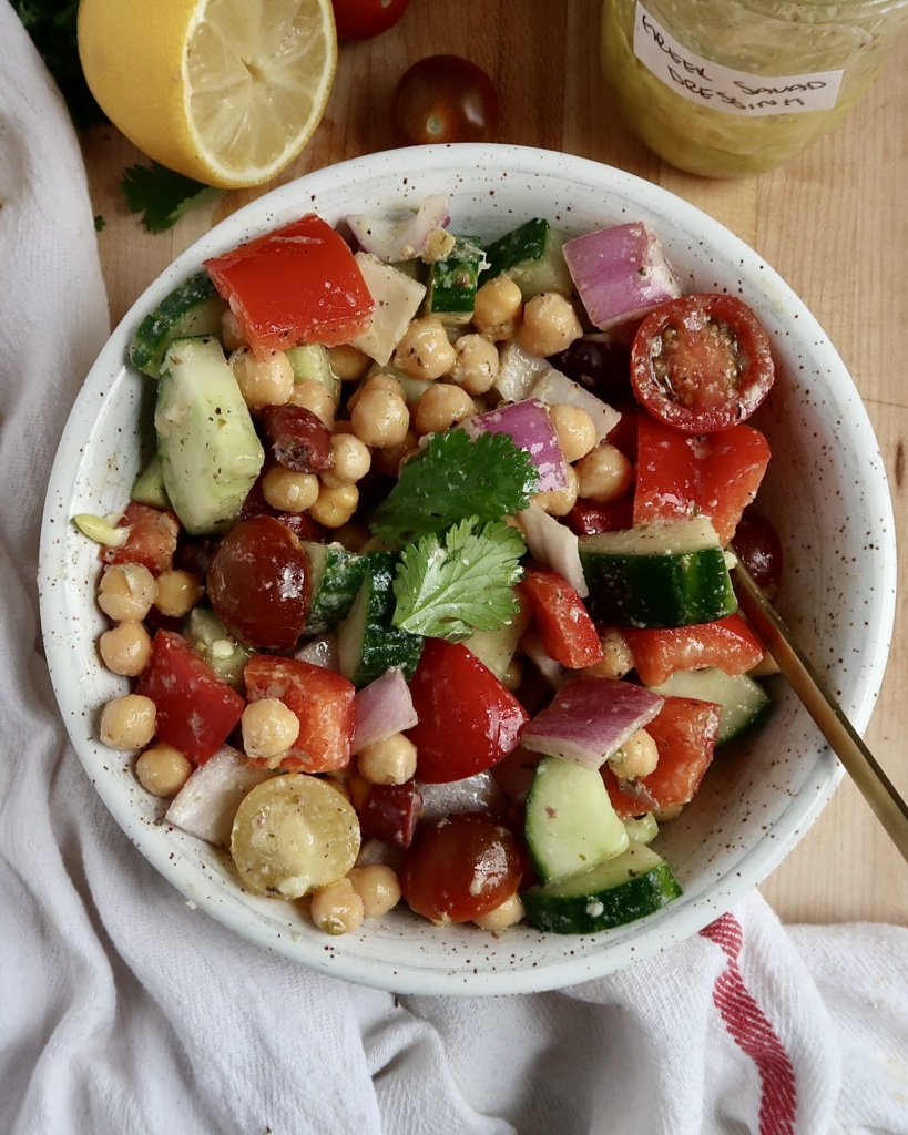 White bowl with chickpeas, tomatoes, cucumber, bell pepper inside and a spoon with a jar labeled 'Greek salad dressing' and a lemon on the side.