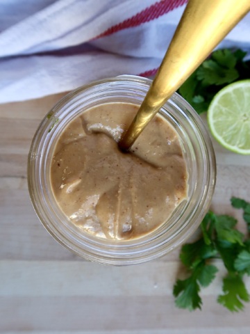 Jar with a creamy peanut sauce inside with a spoon in it and lime to the side.