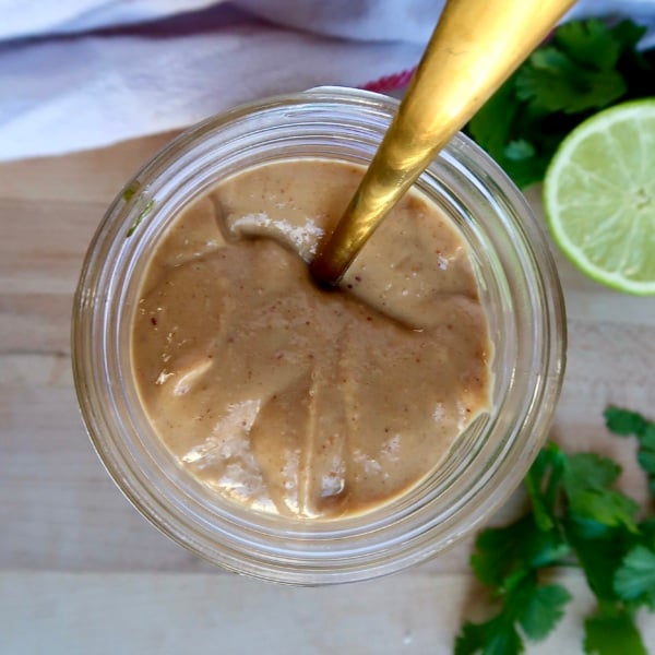 Jar with a creamy peanut sauce inside with a spoon in it and lime to the side.
