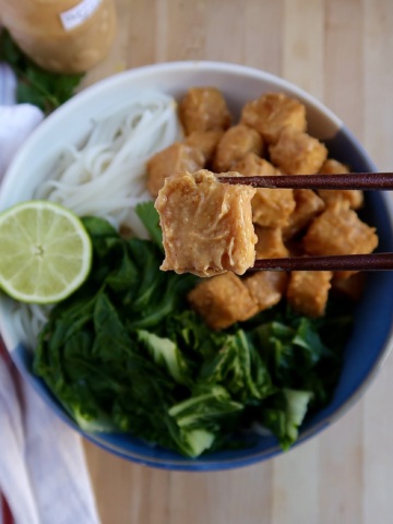 Bowl with rice noodles, bok choy, and peanut tofu with chopsticks holding up a piece of peanut tofu.